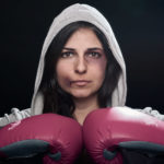 A dramatic portrait of a bruised female boxer wearing boxing gloves and a gray hoodie posing against a dark background