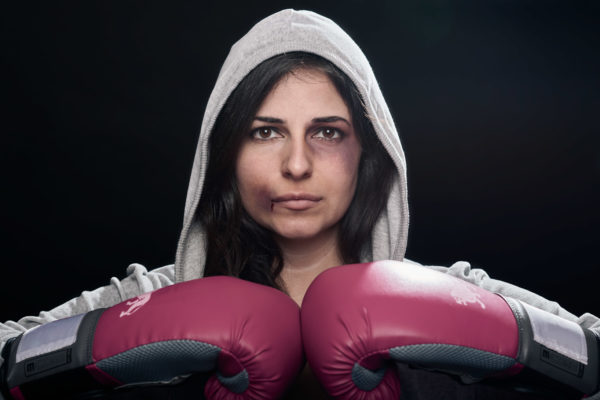A dramatic portrait of a bruised female boxer wearing boxing gloves and a gray hoodie posing against a dark background
