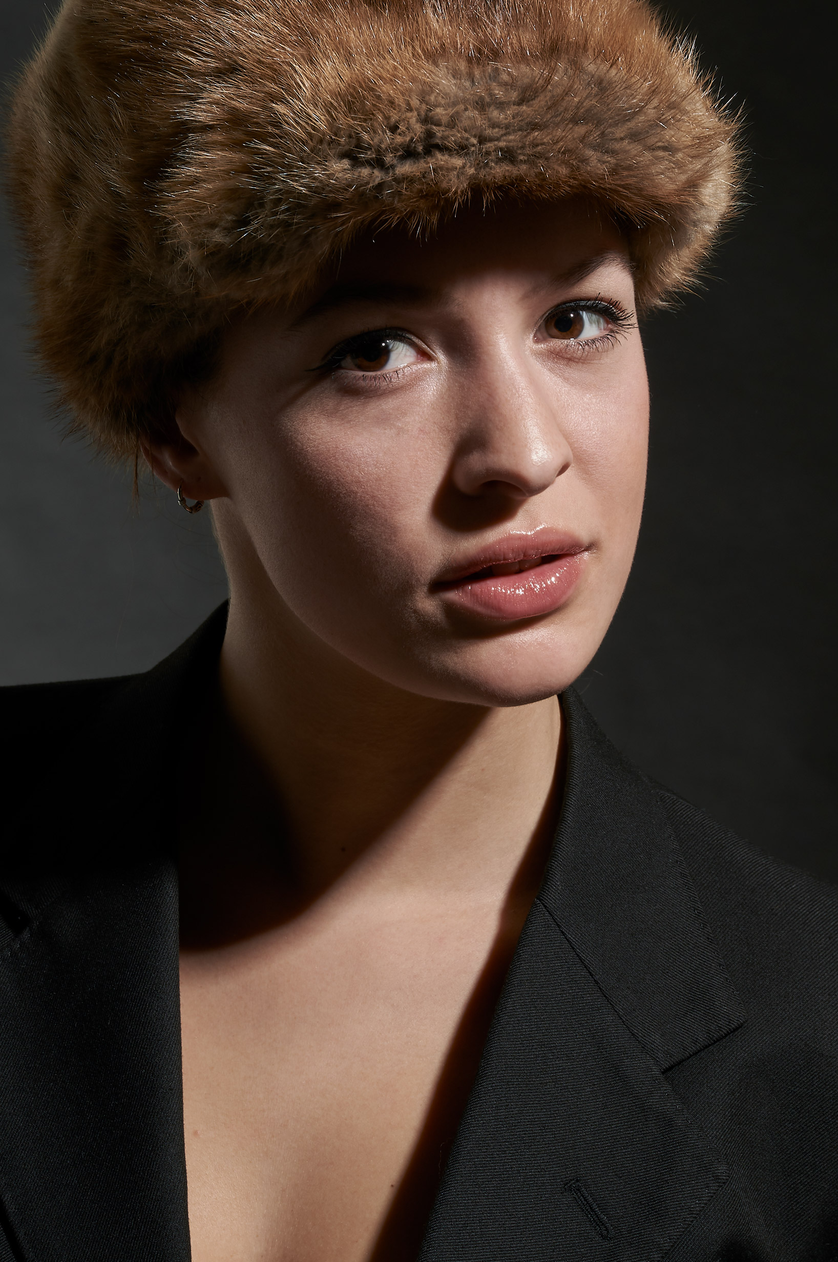 A girl with a winter hat poses for a dramatic portrait wearing a black suit jacket and a brown fur hat