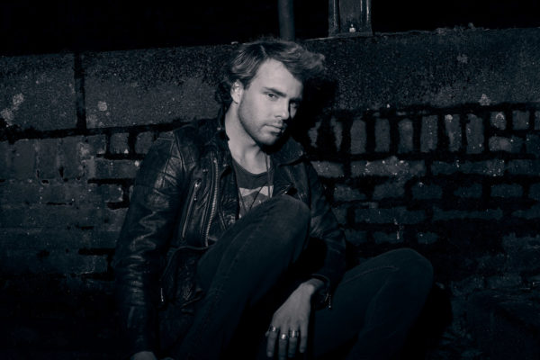 Cinematic portrait of a suspicious man hiding behind a brick wall at night. The man is wearing a black leather jacket as he crouches and hides in the darkness.